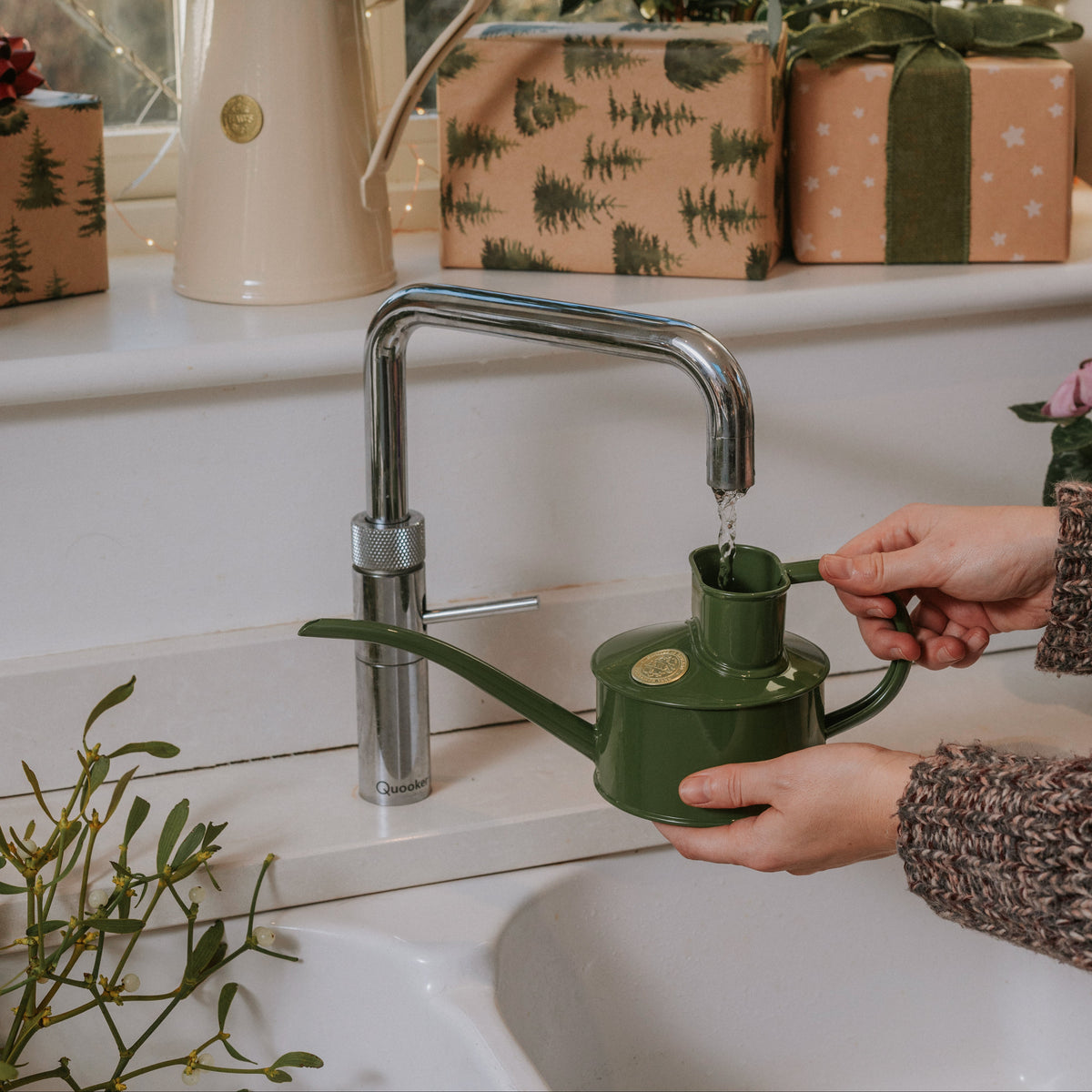 A person's hands fill a dark green metal watering can with water from a chrome faucet in a kitchen sink, with a window, potted plants, and wrapped gift boxes in the background.