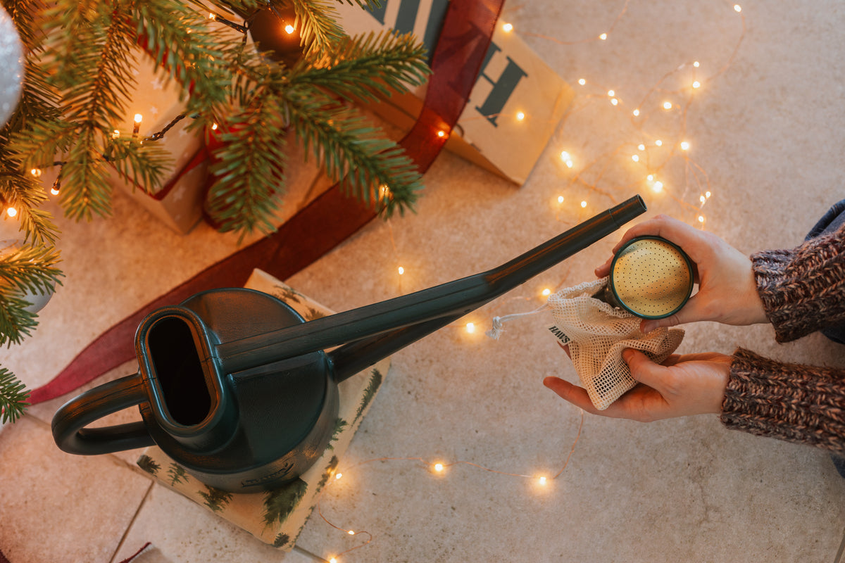 A dark green ceramic watering can sits on a stone floor next to a person's hands holding a small glass jar with a yellow sponge, surrounded by warm fairy lights, pine branches, and wrapped gifts.